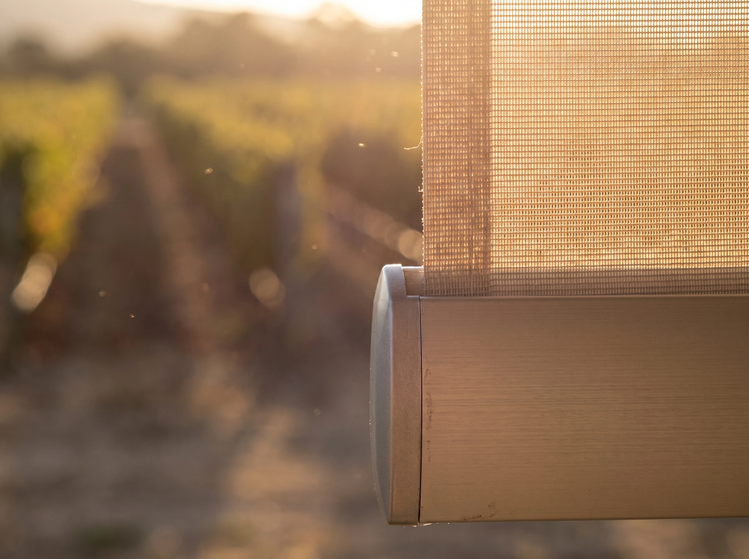 Close-up of sunscreen mesh fabric weave and roller-tube bracket hardware backlit by golden light, vineyard rows out of focus behind